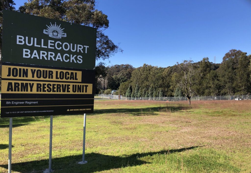Sign reading "Bullecourt Barracks" and "Join Your Local Army Reserve Unit" stands on a grassy area with trees and a clear blue sky in the background. A fence is visible near the wooded area.