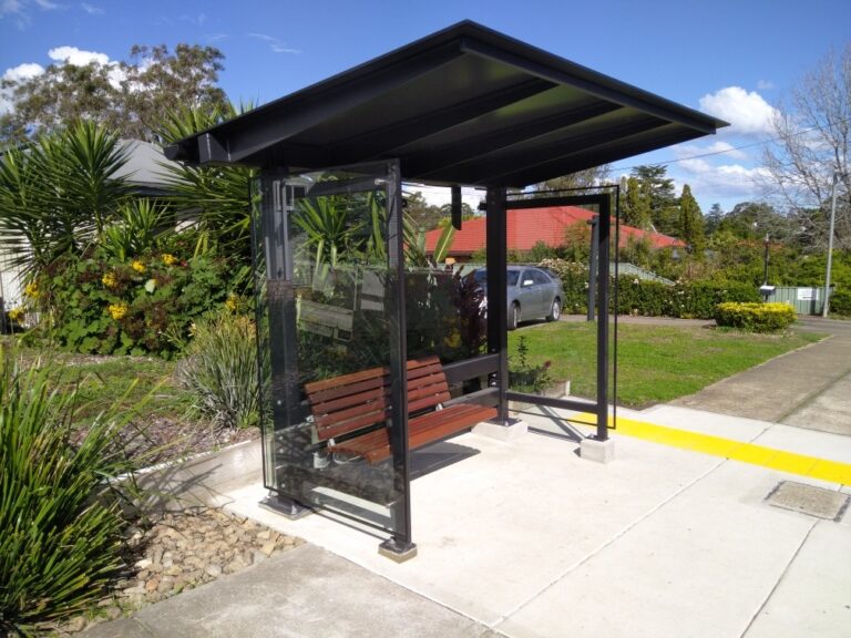 A modern bus stop with a wooden bench and glass panels, surrounded by greenery and plants. A concrete path leads to the shelter, and a yellow tactile strip marks the edge of the sidewalk. The sky is clear and sunny.