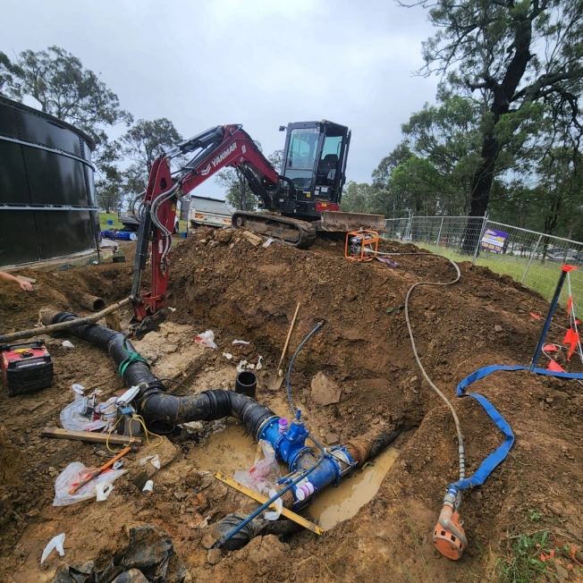 A construction site with a small excavator near a large hole in the ground. Exposed pipes and plumbing materials are visible in the trench. Surrounding the site are trees, construction barriers, and equipment under a cloudy sky.