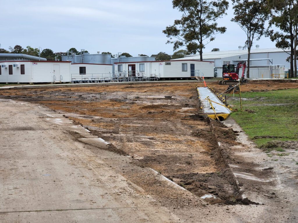 A construction site with cleared soil and a newly paved area beside temporary office trailers. A small red excavator is parked on the right, and caution tape is visible along the edge of the pavement. Trees and overcast skies are in the background.