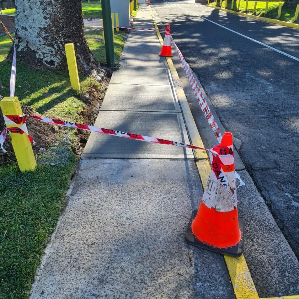 Sidewalk section blocked off with caution tape and orange cones. The area appears to be under construction or repair, with yellow posts and a tree nearby. A road runs parallel to the sidewalk.