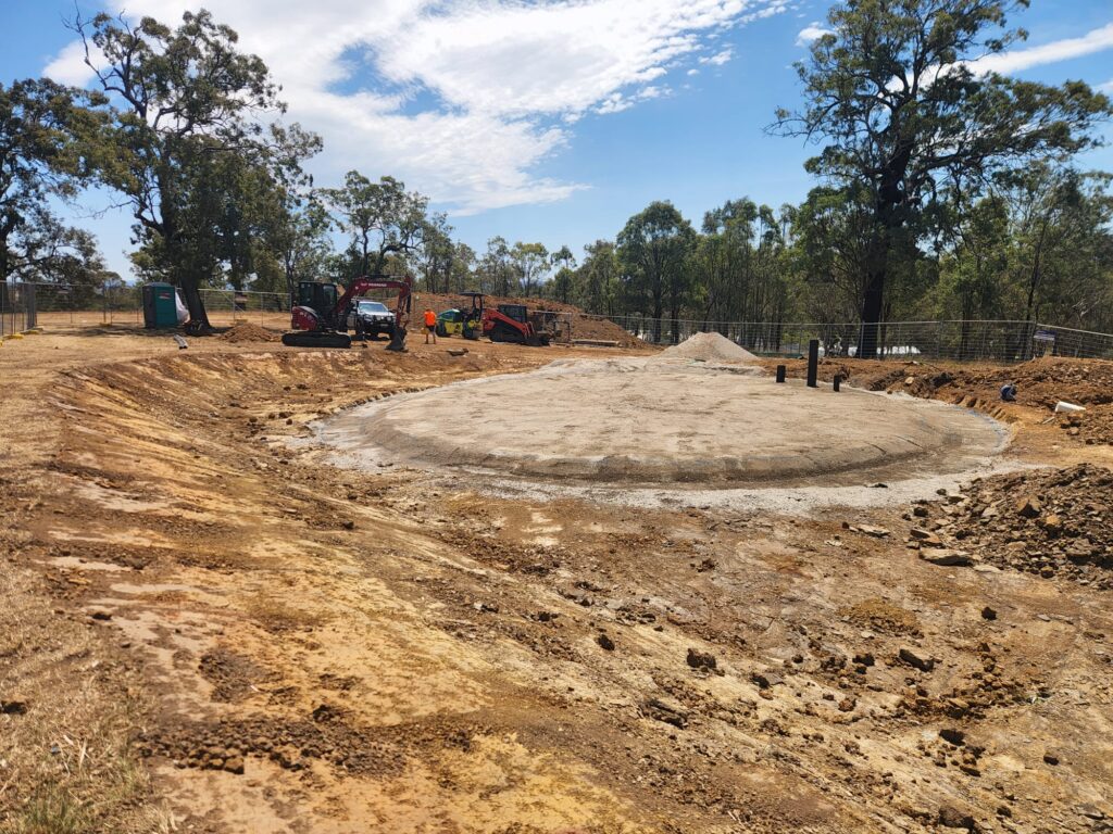 Construction site in a cleared area with a circular foundation and several workers. Excavators and construction equipment are present. Tall trees and fencing surround the site, under a partly cloudy sky.