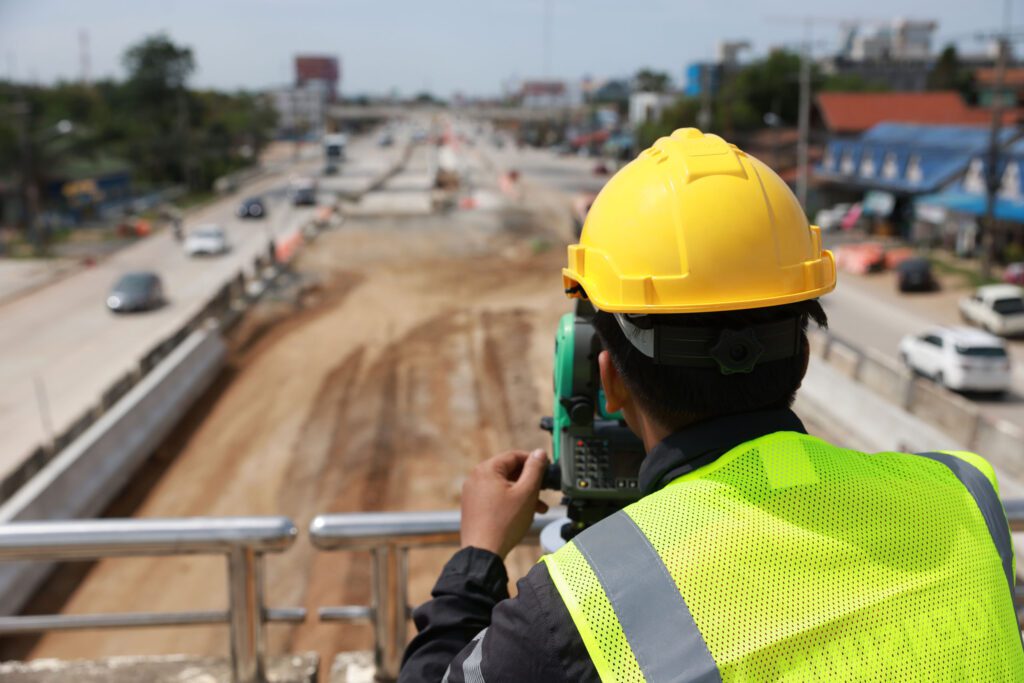 A construction worker wearing a yellow hard hat and green safety vest is using surveying equipment to measure a road construction site. The scene shows traffic moving in the background under a clear sky.