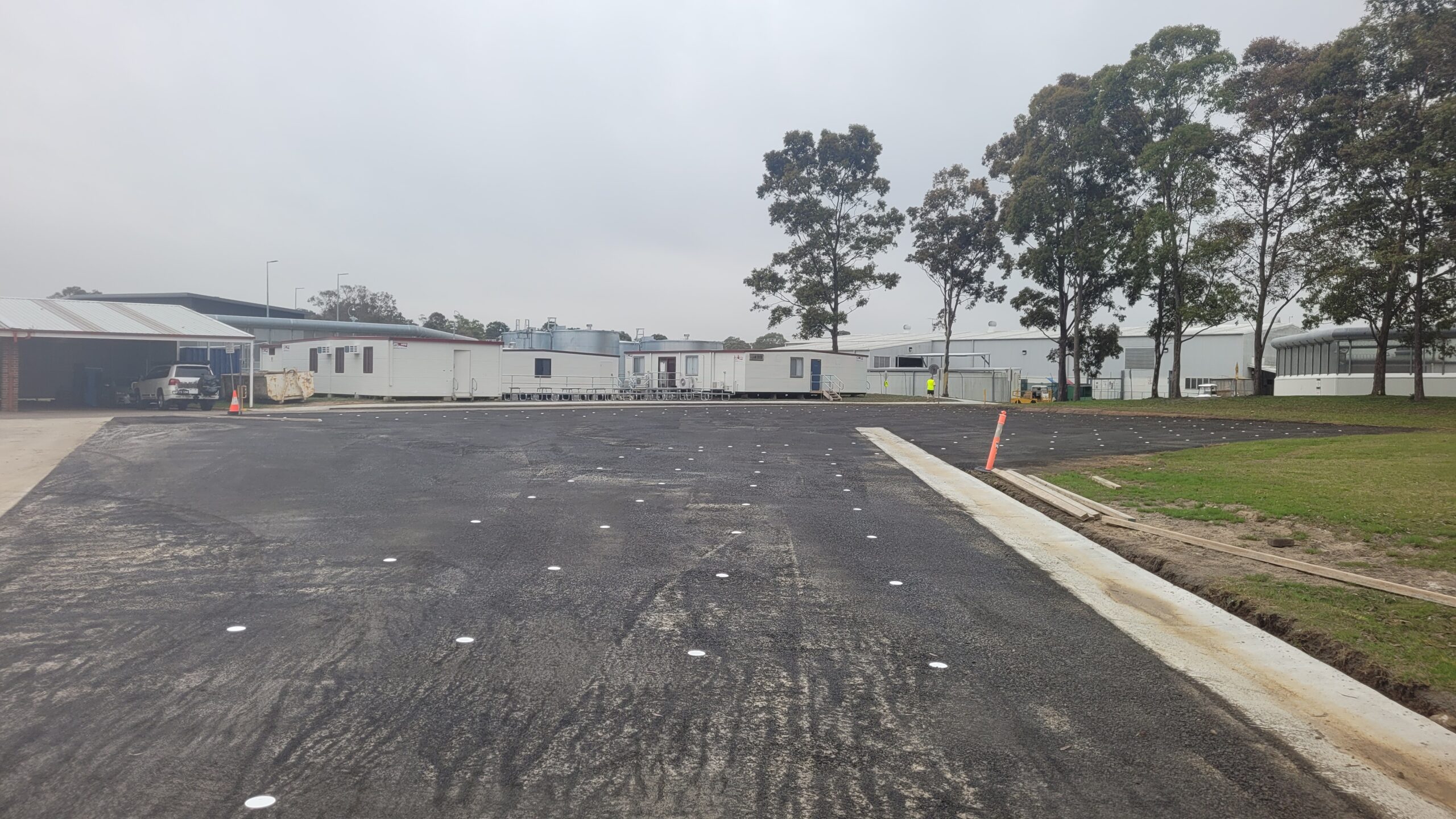 View of a newly paved parking lot with temporary white dots, surrounded by several portable buildings and trees in the background. The sky is overcast, and some construction materials are visible on the left.