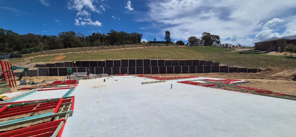Construction site with a concrete foundation and scattered red and green metal frames. Workers in safety gear are on the site. A retaining wall is in the background with green hills and a partly cloudy sky overhead.